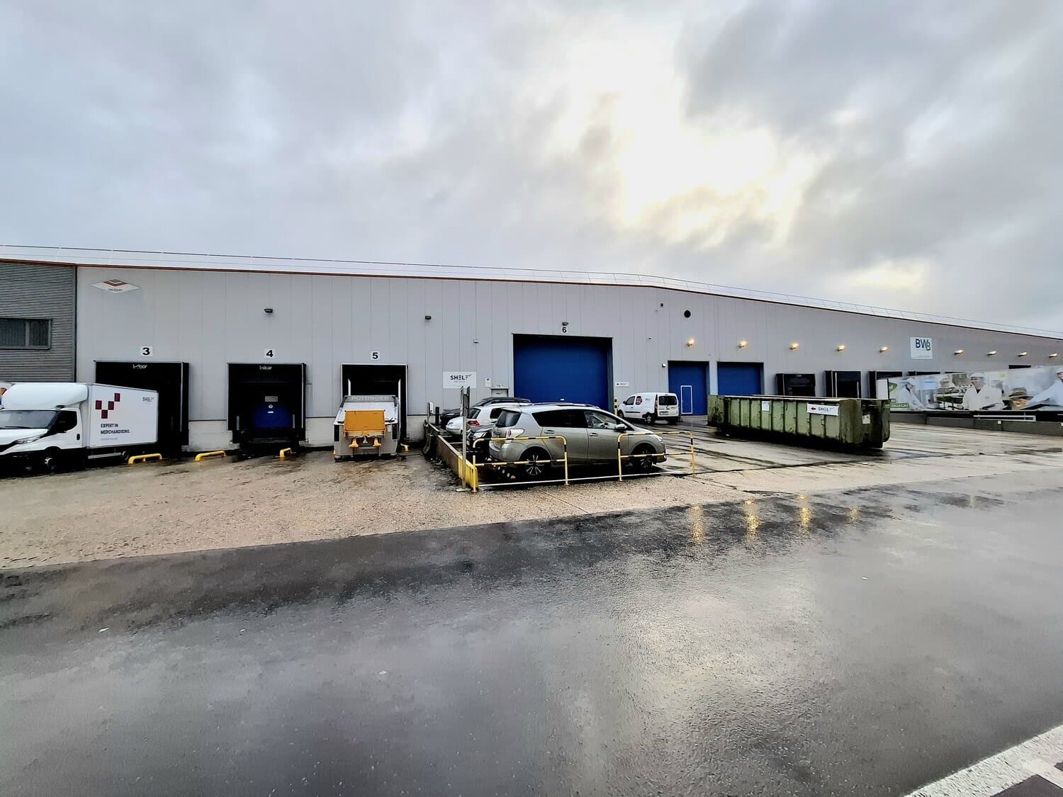 Grey warehouse with numbered loading docks, white truck, car and green skip on a wet parking area