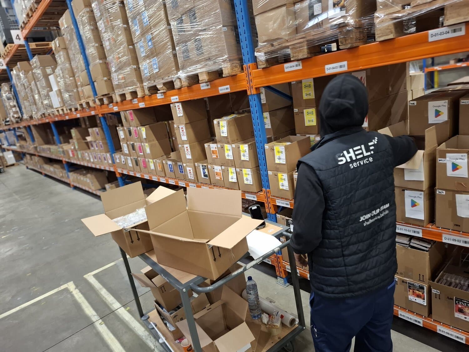 Worker in a Shelf Service vest picking boxes at warehouse shelving, with a cart of open cartons in the foreground