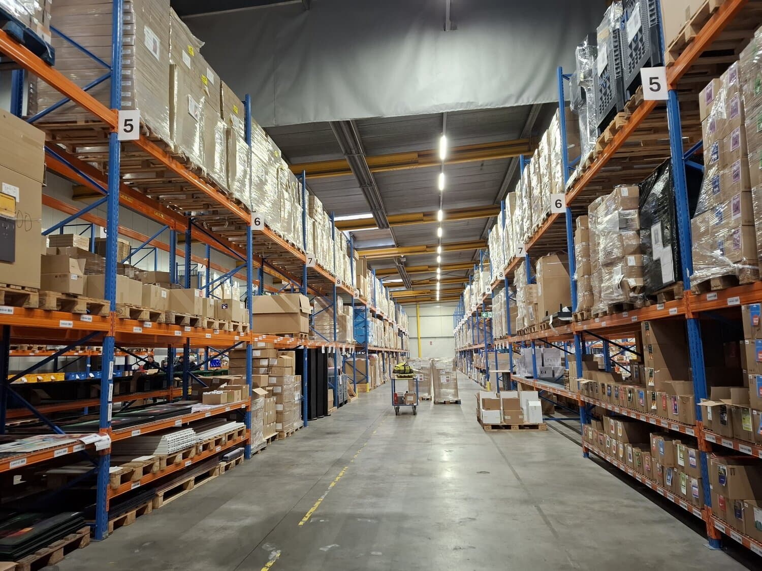 Warehouse aisle between tall metal racks loaded with cardboard boxes and shrink-wrapped pallets, marked with numbers