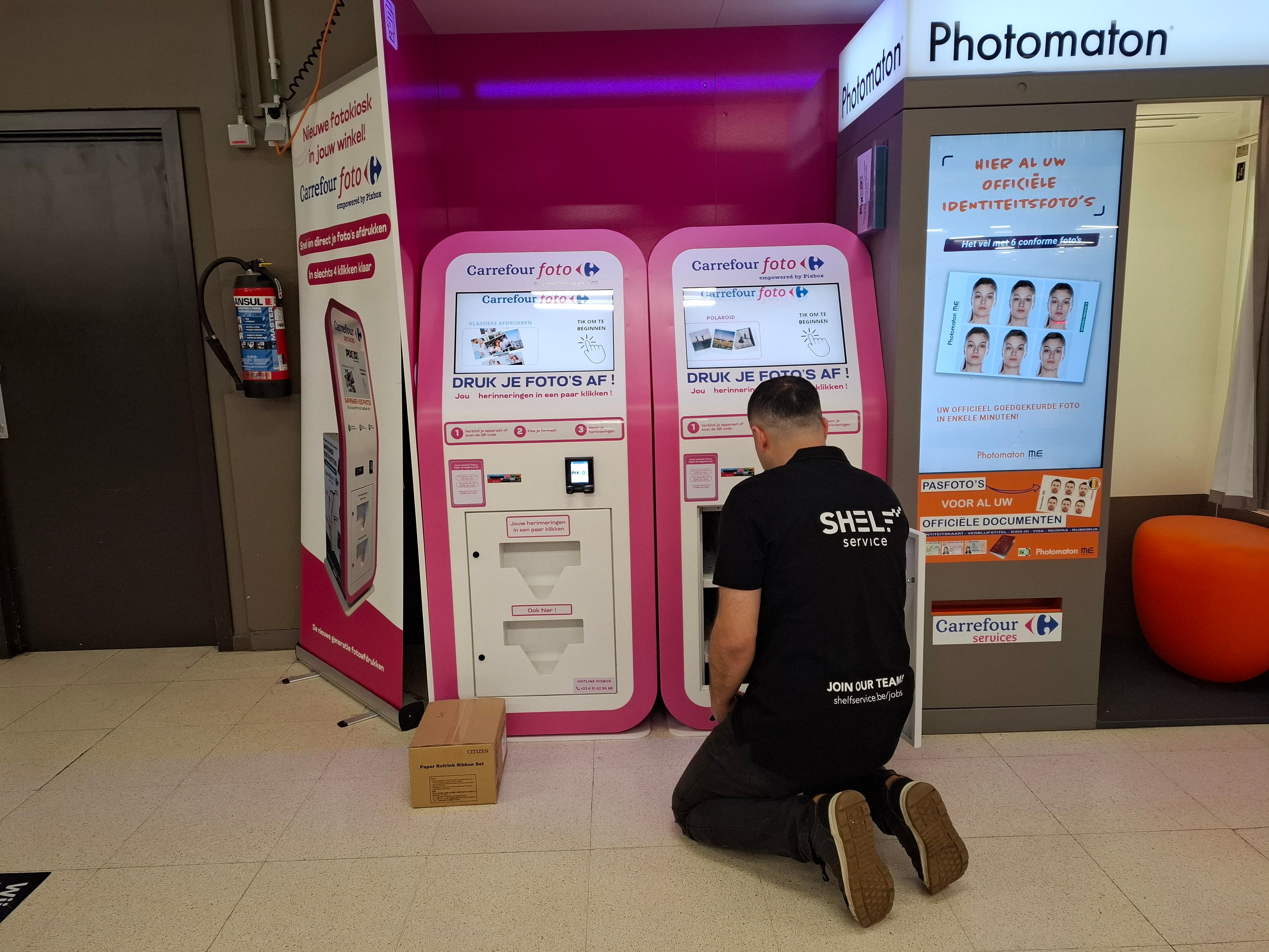 Technician in Shelf Service shirt kneeling in front of two pink Carrefour foto photo kiosks beside a Photomaton booth