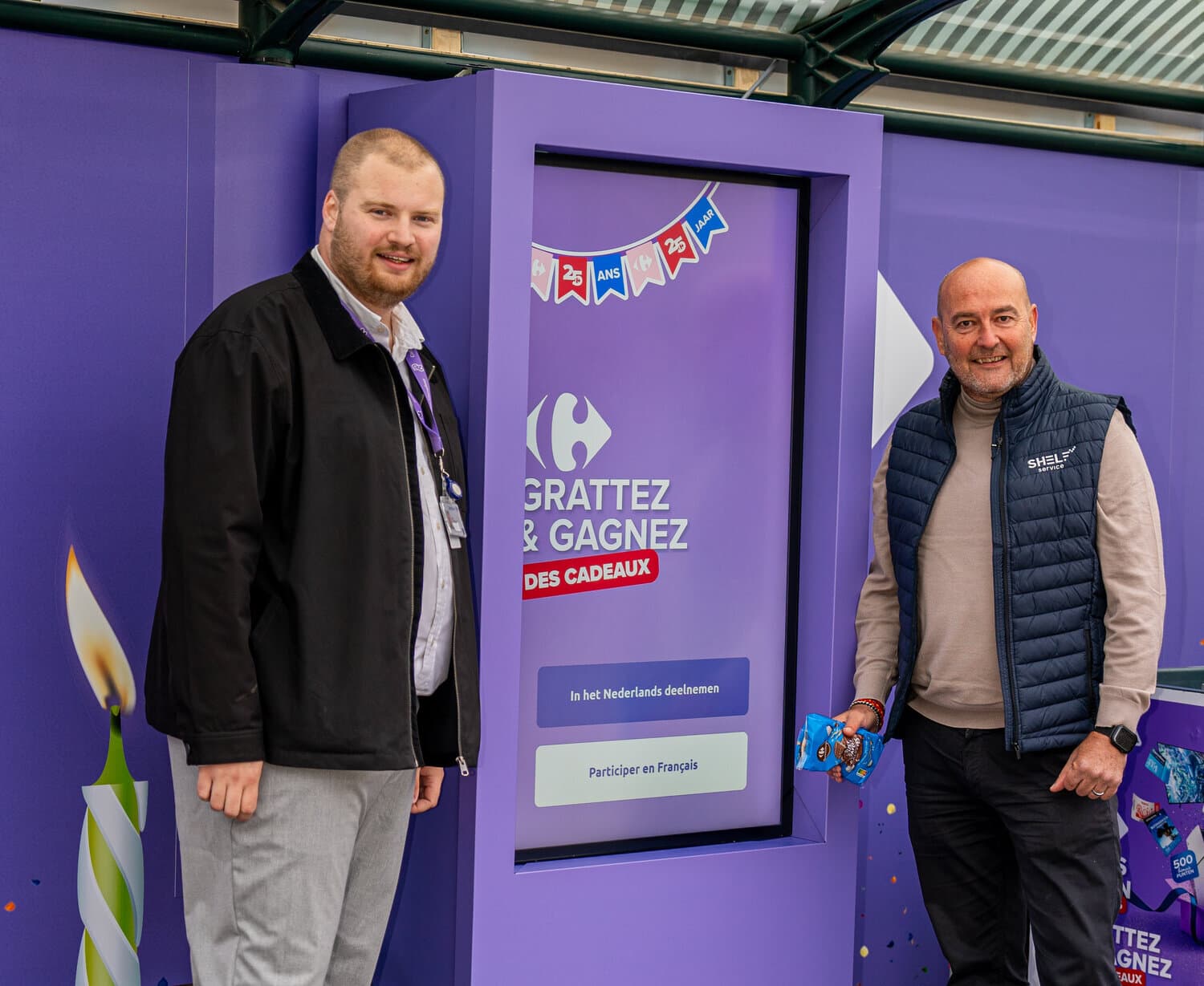 Deux hommes posent devant une borne violette Carrefour affichant « Grattez & Gagnez des cadeaux » et les 25 ans