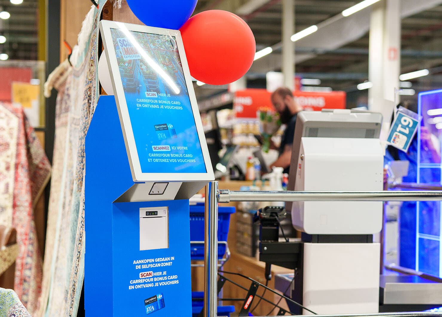 Blauwe Carrefour-zuil met scherm om de Bonus Card te scannen, rode en blauwe ballonnen, kassa's op de achtergrond