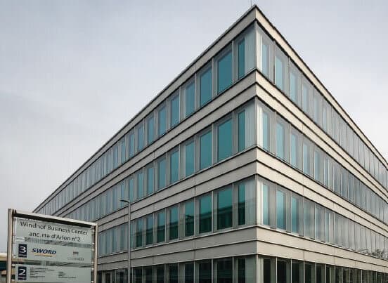 Immeuble de bureaux à façade vitrée, panneau « Windhof Business Center anc. rte d'Arlon n°2 » au premier plan