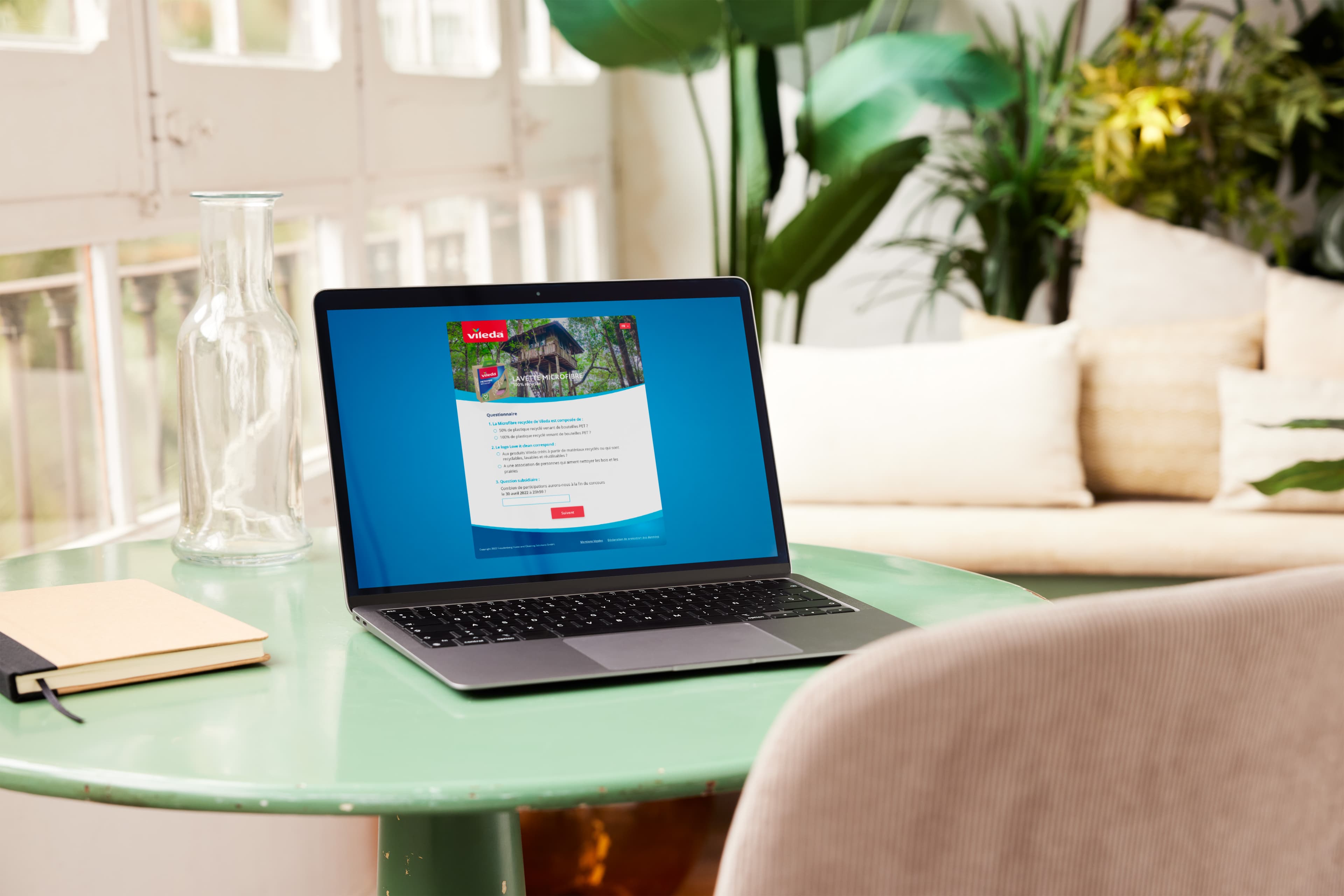 Laptop showing a Vileda questionnaire page on a green table next to a notebook and a glass carafe