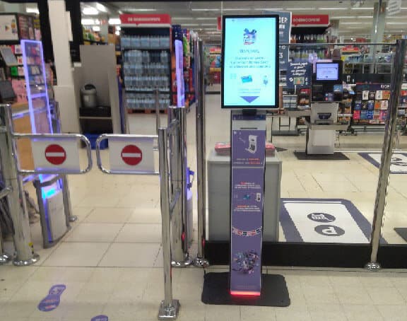 Borne digitale à écran vertical près des portillons d'entrée d'un hypermarché Carrefour, avec cartes cadeaux au fond