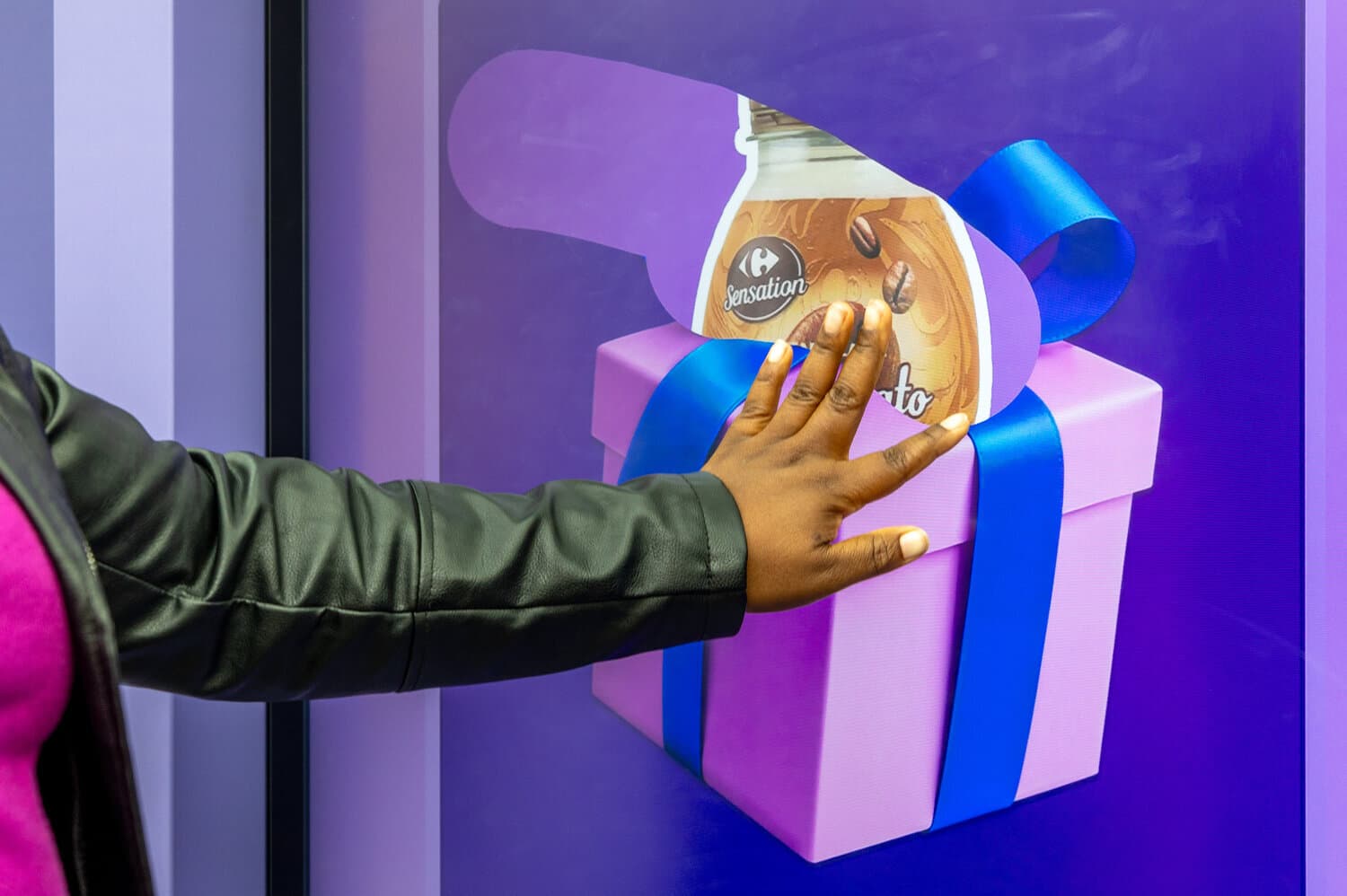 Main posée sur un écran tactile montrant un cadeau violet à ruban bleu et une bouteille Carrefour Sensation