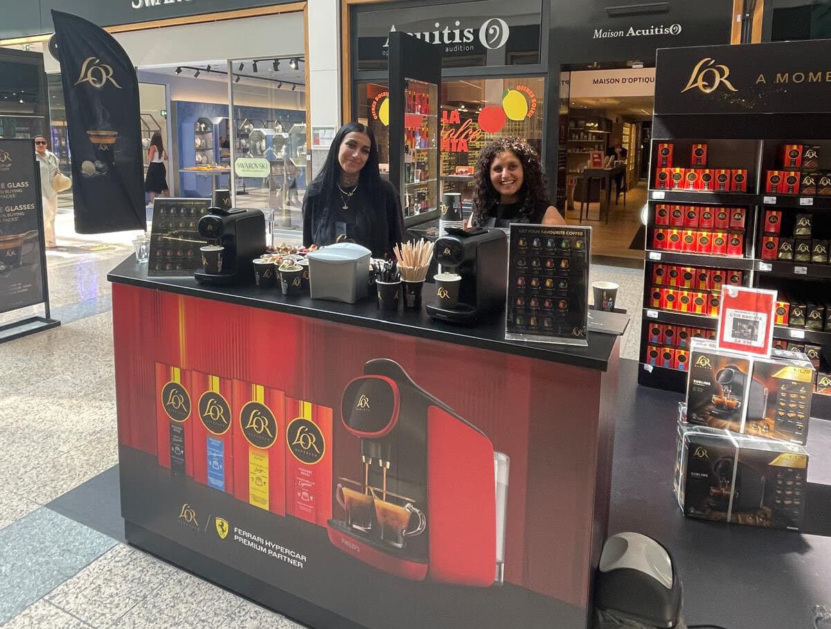 Deux hôtesses derrière un stand de dégustation L'OR avec machines à café, présentoir de capsules et boîtes en galerie commerciale