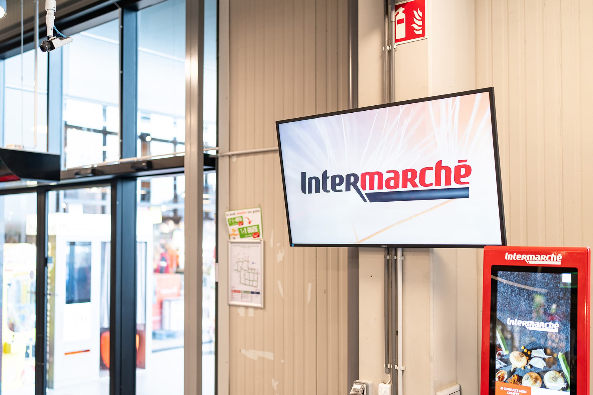 Wall-mounted screen showing Intermarché at a store entrance, next to a red touchscreen kiosk