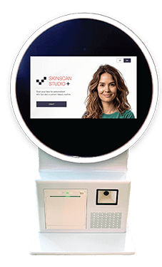 White kiosk with round screen showing "Skinscan Studio+" and a portrait of a smiling woman