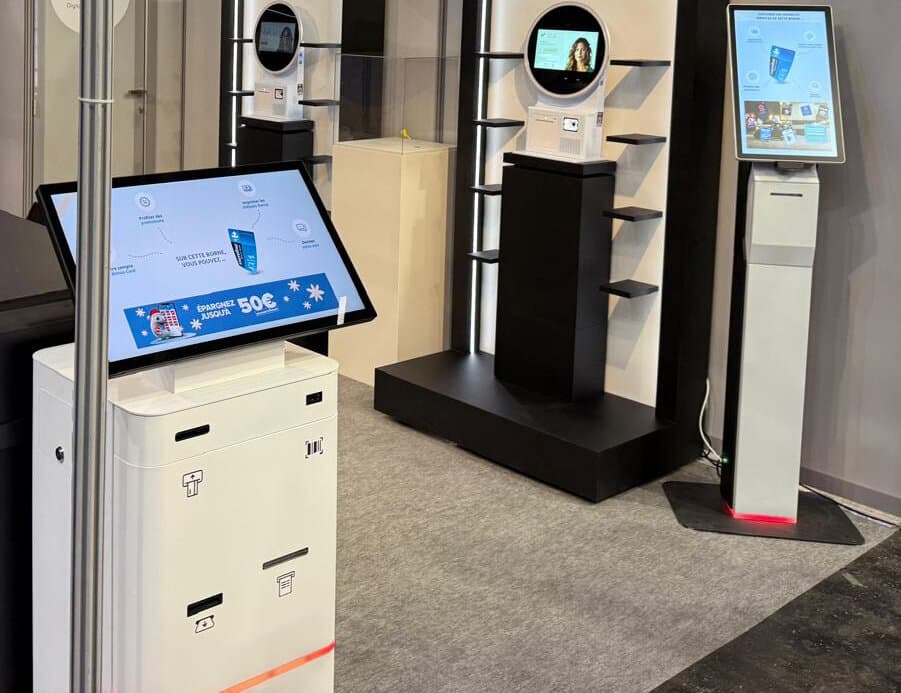 Witte touchscreenzuilen en zwart rek met planken op een stand, scherm met de tekst « Épargnez jusqu'à 50€ »