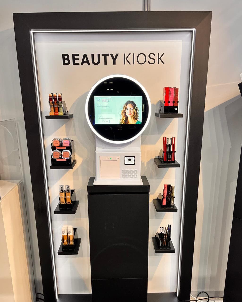 Beauty Kiosk-zuil met rond scherm, omringd door schappen met make-up: foundations, blushers, lippenstiften en mascara's