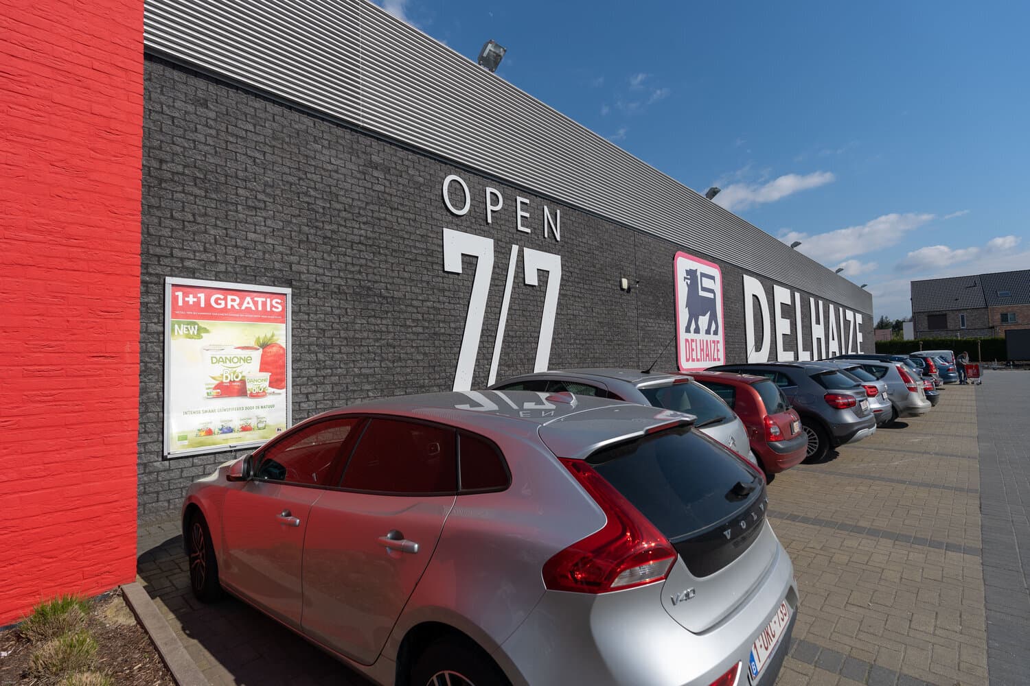 Delhaize store facade with a Danone 1+1 gratis poster and cars parked in the car park