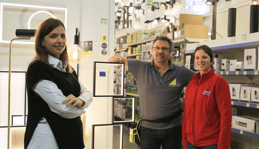 Three people pose in a store lighting aisle among lamps and product boxes, one wearing a red Hubo fleece