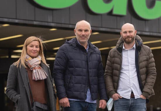 Une femme et deux hommes en vestes d'hiver posent devant l'entrée d'un magasin aux grandes lettres vertes