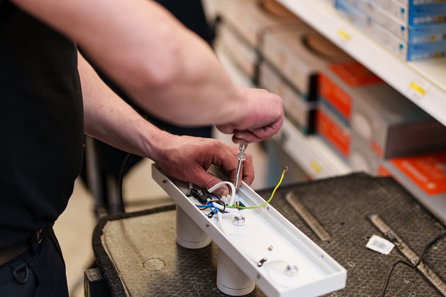 Technician's hands wiring a white light fixture with a screwdriver in front of DIY store shelves