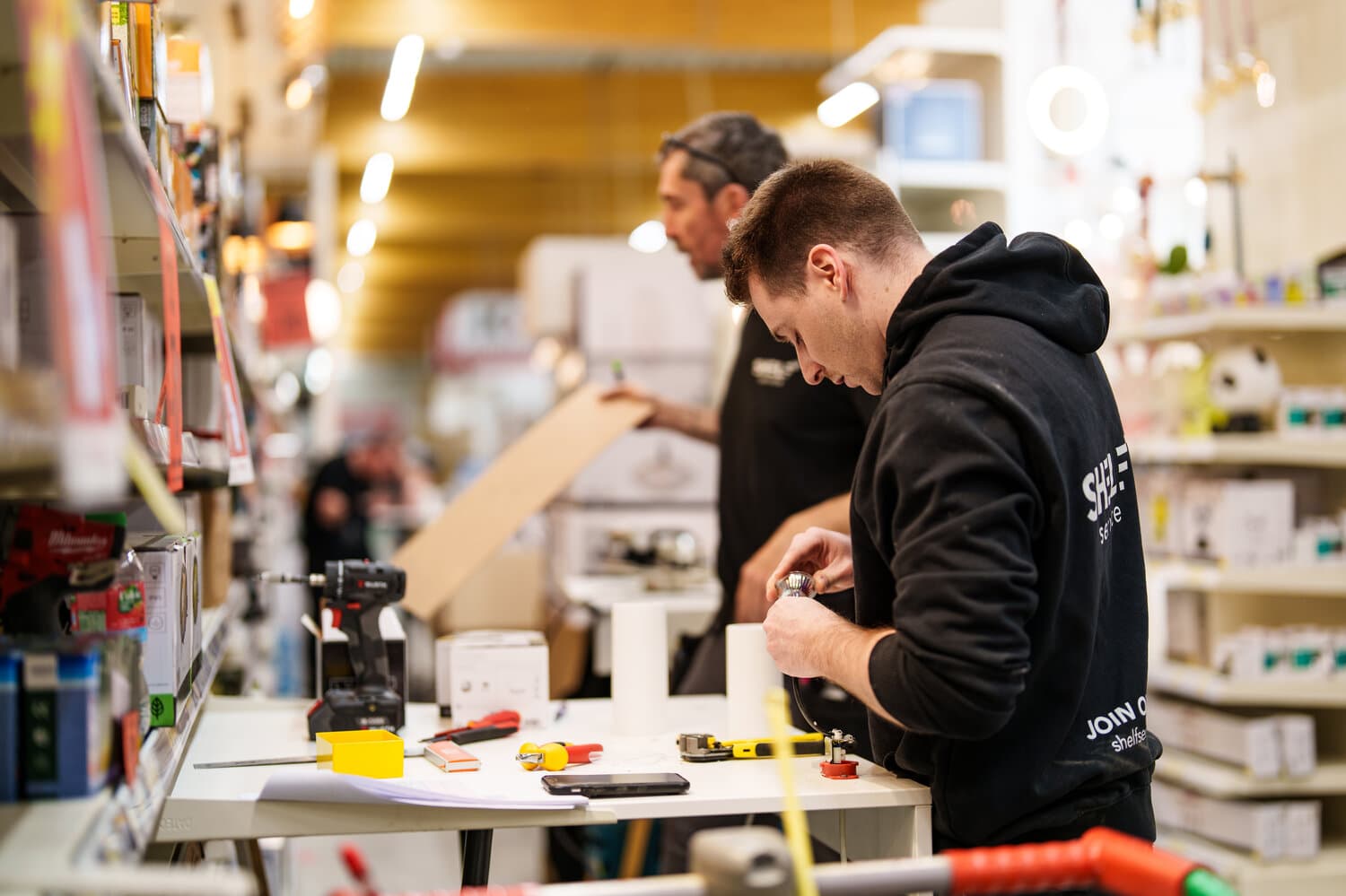 Two merchandisers in black hoodies work on a product at a table in a DIY store