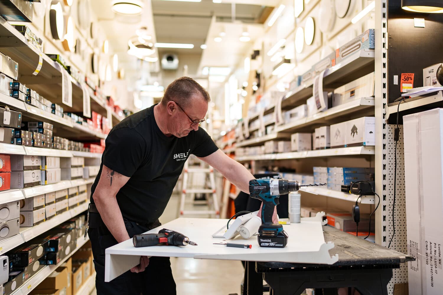 Shelf Service technician drilling a white shelf in the lighting aisle of a DIY store