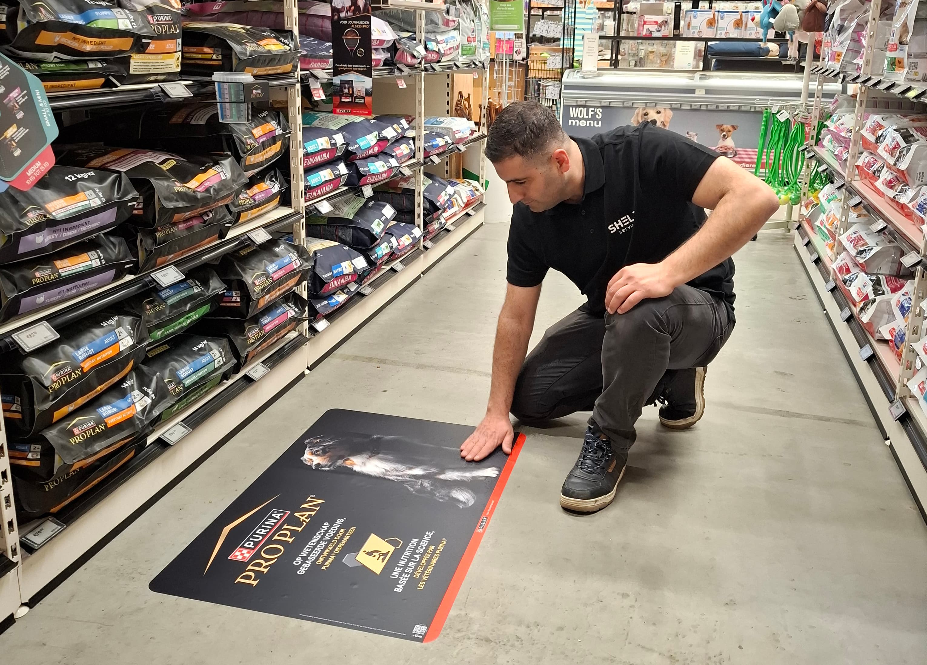 Merchandiser hurkt en legt zijn hand op een Purina Pro Plan-vloersticker in een schap met hondenvoeding