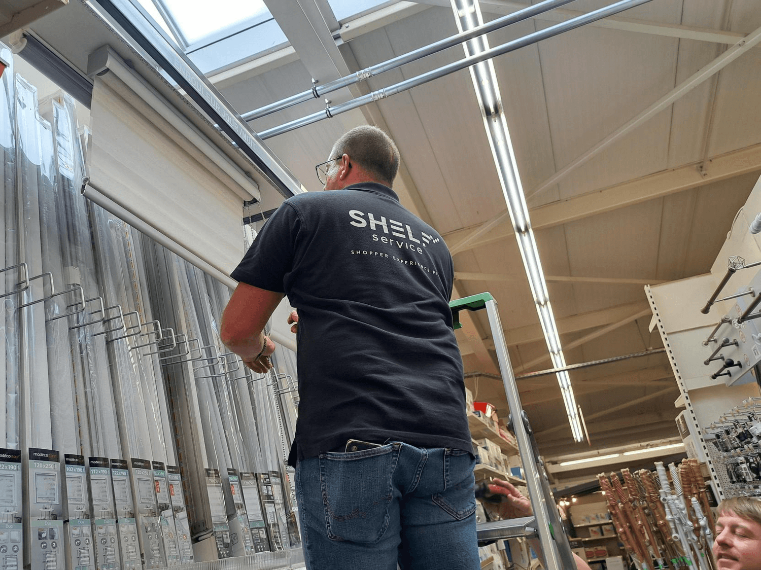 Technicus in Shelf Service-poloshirt plaatst rolgordijnen in een wandschap van een doe-het-zelfzaak