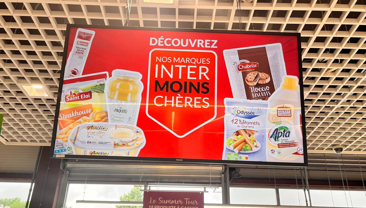 Écran suspendu en magasin annonçant « Découvrez nos marques Inter Moins Chères » avec des emballages de produits alimentaires