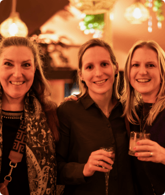 Three smiling women pose side by side holding drinks in a warmly lit venue