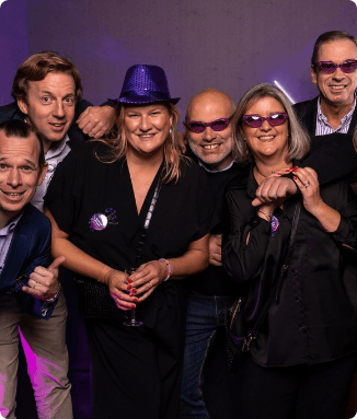 Group of six smiling people in dark clothes, some with purple glasses and a sequin hat, against a purple background