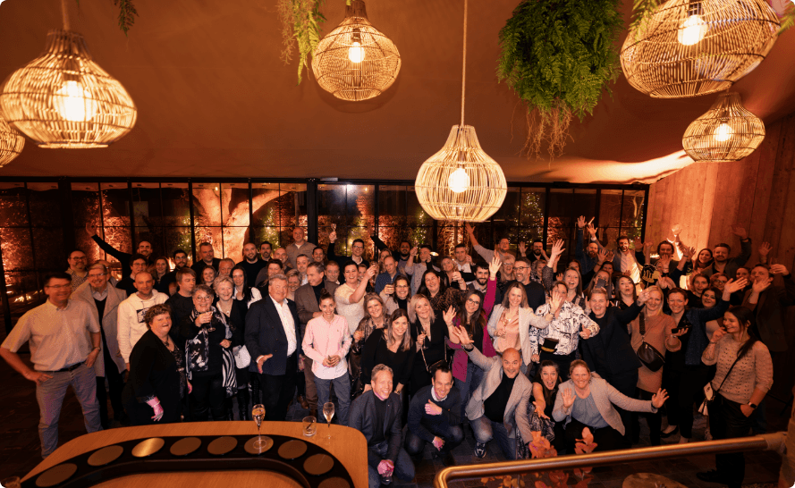 Large group of smiling people raising their arms, seen from above, beneath rattan pendant lamps in a hall