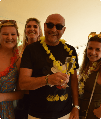 Four smiling people wearing flower garlands, a man in the center holding a glass of champagne