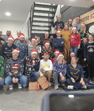 Group of employees in Christmas sweaters and Santa hats posing in front of a staircase with gifts
