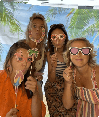 Four women pose in front of a palm tree backdrop holding photo booth props on sticks: glasses, parrot, palm tree